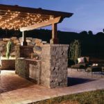 An outdoor kitchen with arbor and lights at night by Backyard Paradise.